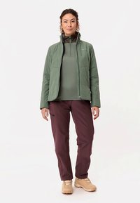 Groene geïsoleerde jas over een groen topje, gecombineerd met bordeauxrode broek en beige schoenen. Voorzien van ritssluitingen en zijzakken.