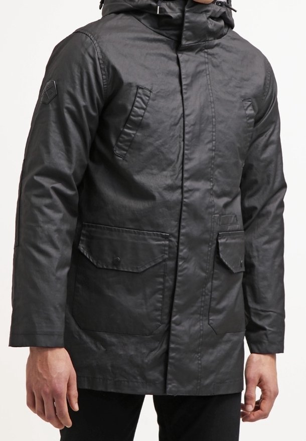 Veste en coton ciré noir à capuche avec fermeture éclair devant, poches à rabat et poches verticales sur la poitrine portée par une personne.