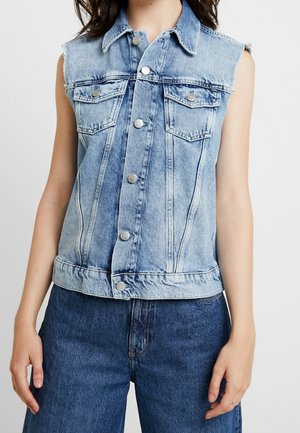 Lys blå denimvest uden ærmer med knaplukning og brystlommer med klap, båret med mørkeblå denimbukser.