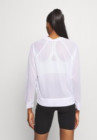 Weißes Mesh-Oberteil mit langen Ärmeln in lockerer Passform, ausgestattet mit einem runden Ausschnitt und sichtbarem Lagenlook über einem Sport-BH, kombiniert mit schwarzen Shorts.