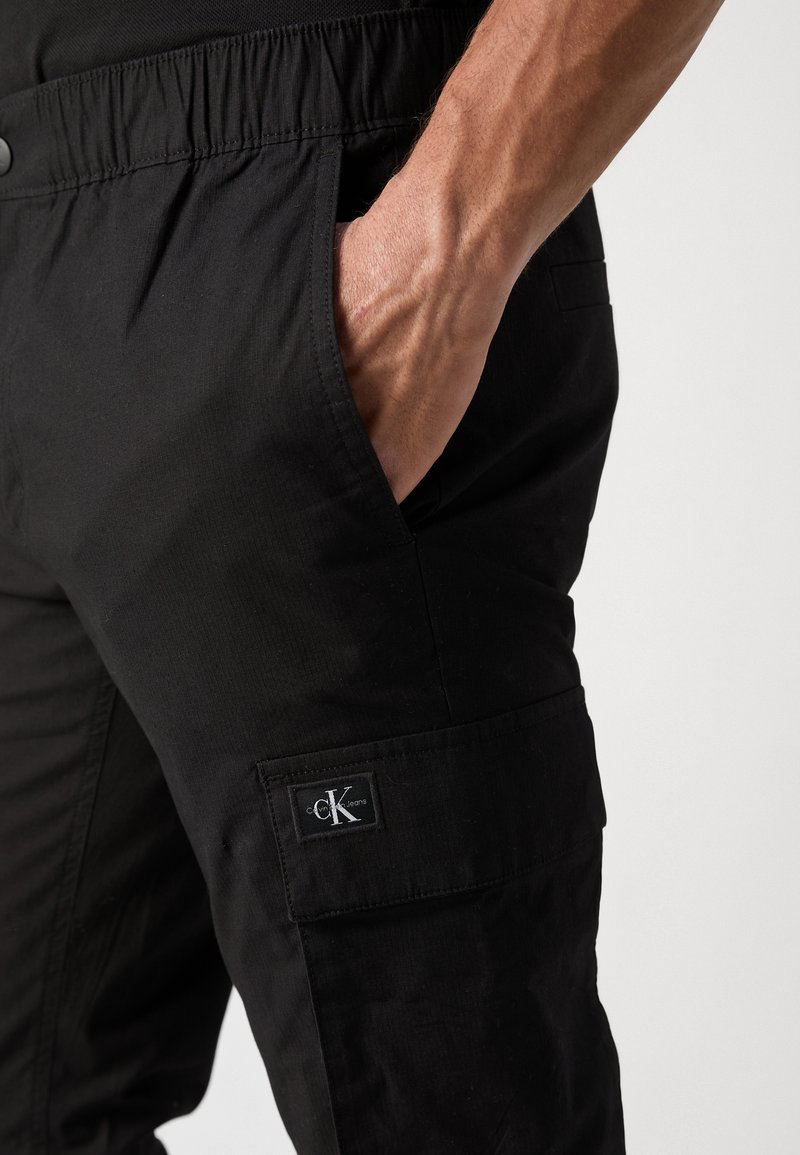 Pantaloni cargo neri con vita elastica, dotati di tasche laterali e un piccolo logo ricamato sulla coscia sinistra. Il materiale sembra liscio e resistente.