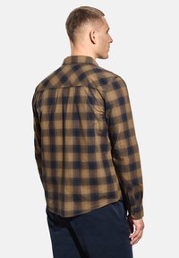 Muž v hnědém a námořnickém kostkovaném dlouhém rukávu, stojící mírně otočený doprava na jednobodovém světle.