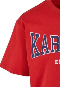 Sarkans kokvilnas T-krekls ar izteiksmīgu zilu printētu "KAR" dizainu ar baltām akcentiem, ar apaļu dekoltē un īsām piedurknēm.