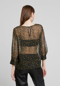 Musta läpinäkyvä paita, jossa on kultaisia ja oransseja kukkakuoseja, pitkät hihat, avonainen selkäosa ja väljä malli.