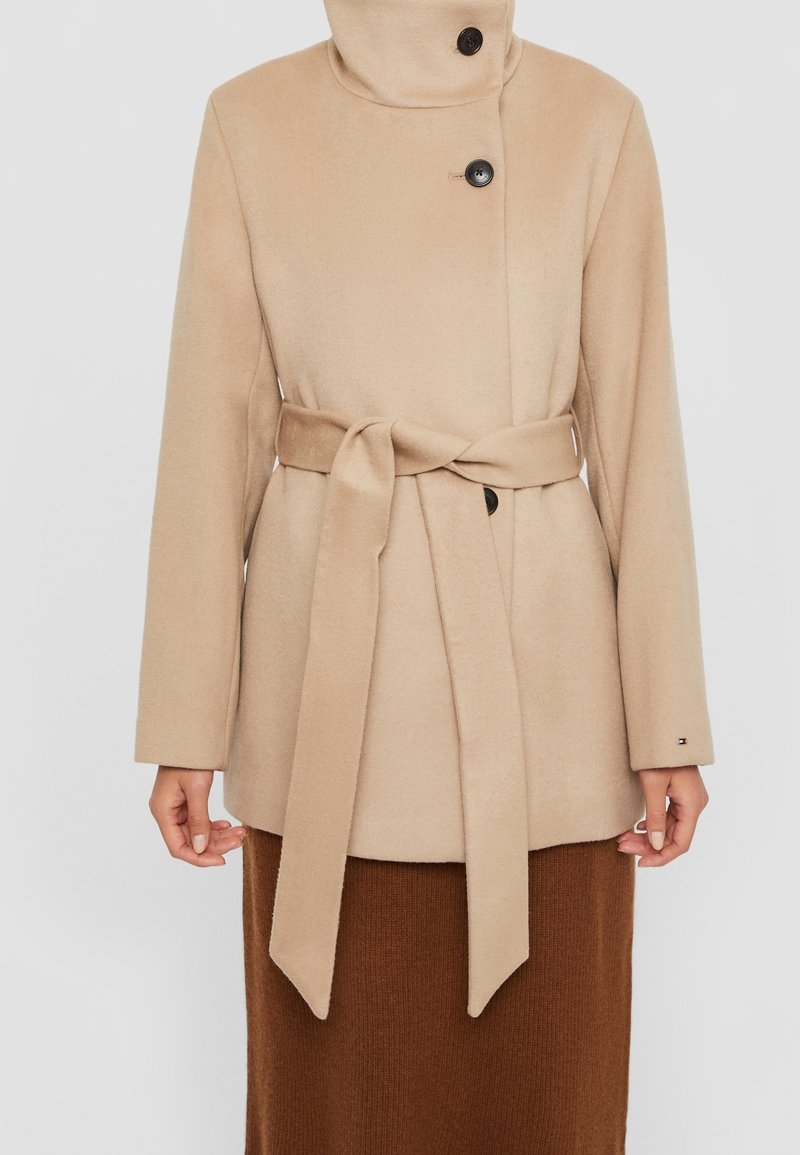 Beige jas van een wolmix met een hoge kraag, een dubbele knoopsluiting en een ceintuur om te strikken. Heeft lange mouwen en een gladde, zachte textuur.