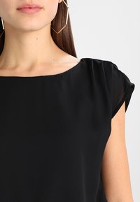 Blusa negra con mangas cortas y fruncidas, escote redondeado y textura de tela suave. Diseño simple sin patrones ni acentos visibles.