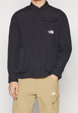 Mand iført en sort The North Face jakke og beige bukser med The North Face-logo på brystet og låret.