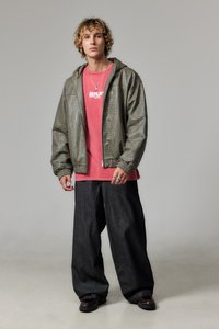 Chaqueta con capucha de tela caqui con estampado, camiseta gráfica roja debajo, pantalones de mezclilla oscuros y holgados, y zapatos negros. Se ven detalles en joyería plateada.