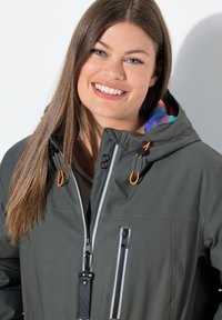 Chaqueta impermeable de color verde oscuro con acentos naranjas, múltiples bolsillos con cremallera y un forro de capucha con un patrón colorido. Textura suave y diseño ajustado.