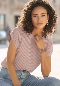Kurzärmliges, geripptes mauve Oberteil, kombiniert mit hellblauen Jeans, ergänzt durch ein mehrsträngiges, metallisches Armband und hängende Ohrringe.