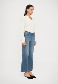 Mulher vestindo um suéter de malha creme com mangas largas, jeans retos azuis de cintura alta e sapatos de salto baixo pretos com bico fino, de pé de lado.