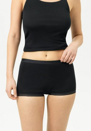 Frau trägt schwarzen ärmellosen Croptop und schwarze enganliegende Shorts und steht vor einem schlichten hellen Hintergrund.
