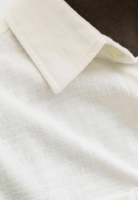 Chemise beige clair avec un tissu texturé à tissage subtil, col pointu et un seul bouton décoratif sur le col.