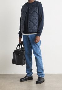 Navy-gestepptes Puffer-Westen über einem blauen Sweatshirt, kombiniert mit hellblauen Jeans und einer schwarzen Lederhandtasche mit Reißverschluss. Schwarze Knöchelstiefel runden den Look ab.