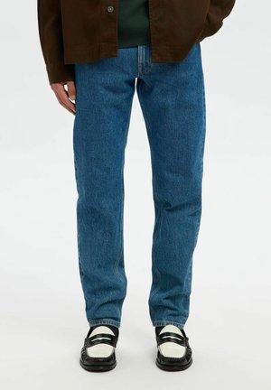 Person wearing blue straight-leg jeans, black and white loafers, with part of a brown jacket and green shirt visible.