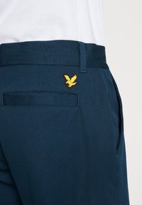 Marinblå byxor i slät tyg, med en bakficka som har en gul fågel-emblem. Midjan är platt med en subtil textur.