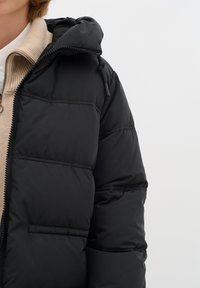Svart pufferjacka med vadderad design, som har huva och dragkedjestängning. Tyget är glatt med en lätt glans och synliga sömmar.