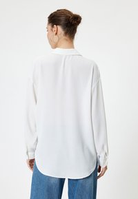 Weißes Bluse mit langen Ärmeln und Kragen, lockere Passform und gerundetem Saum. Glatte Textur mit knöpfbaren Manschetten, kombiniert mit blauen, hoch taillierten Jeans.