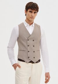 Jasnobrązowa kamizelka w wzór houndstooth z podwójnym zapięciem, brązowymi guzikami i dwoma przednimi kieszeniami, noszona na białej koszuli i beżowych spodniach.