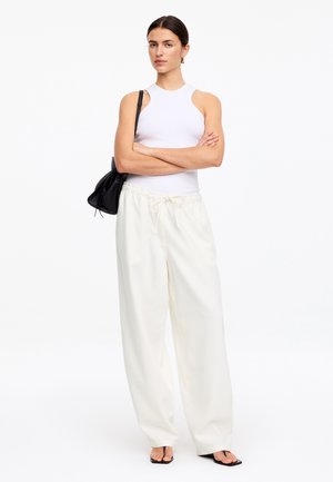 Žena stojí so založenými rukami, má na sebe biely tielko bez rukávov, voľné biele nohavice so sťahovacou šnúrkou, čiernu kabelku cez rameno a čierne sandále s otvorenou špičkou.