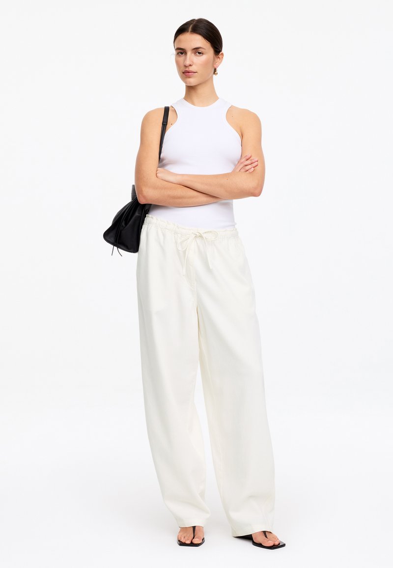 Femme debout, bras croisés, portant un haut blanc sans manches, un pantalon blanc ample avec cordon, un sac à épaule noir et des sandales noires ouvertes au niveau des orteils.