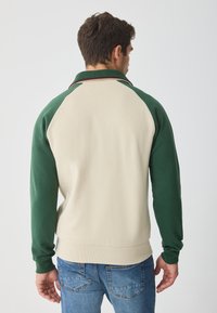 Jersey crema y verde con mangas raglán, que cuenta con un collar y puños acanalados. Hecho de material suave, con detalles de costuras.