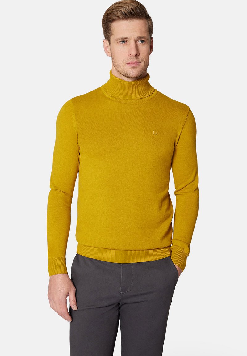 Żółty sweter z golfem wykonany na drutach, o dopasowanym kroju oraz ściągaczach przy mankietach i na dolnej krawędzi. Posiada małe logo na piersi. Noszony z szarymi spodniami.