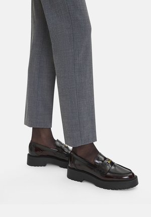 Schwarze Loafer aus Lackleder mit einer klobigen Sohle und dekorativem Metall vorne, kombiniert mit grauen, maßgeschneiderten Hosen.