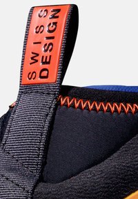 Czarna teksturowana tkanina z pomarańczowym przeszyciem i czerwoną metką z wytłoczonym napisem "SWISS DESIGN" w kolorze czarnym, przymocowaną do górnej części buta.