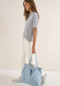 Femme marchant en chemise à manches courtes rayée, pantalon crème, baskets argentées, portant un grand sac cabas bleu clair.