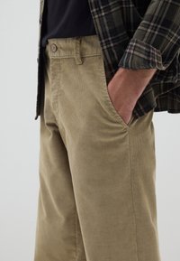 Pantaloni in corduroy color cannella con tessuto texture, tasche laterali e vita aderente. Abbinati a una camicia a quadri scura.