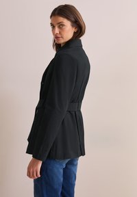Černý přizpůsobený blazer s opaskem, strukturovanými rameny, dvěma knoflíky a skládáním na zadní straně, v kombinaci s modrými džíny. Hladká struktura tkaniny.