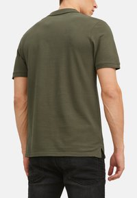Olivgrünes Polohemd, kurze Ärmel, weiche Textur, gerader Saum mit Seitenschlitzen, getragen mit dunklen Jeans, schlichte Gestaltung ohne Verzierungen.