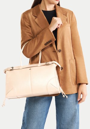 Žena ve svetrovém blejzru barvy velblouda a modrých džínách držící velkou béžovou koženou kabelku za uchy po svém boku.