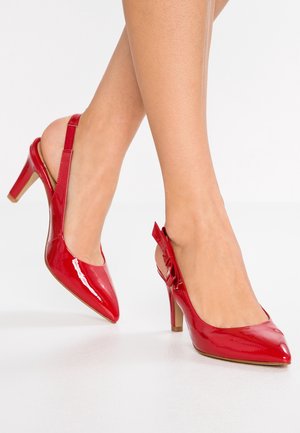 Vrouw draagt glanzende rode slingback hoge hakken met spitse neuzen en een kleine decoratieve strik aan de zijkant.