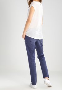 Vrouw in een witte blouse met korte mouwen, marineblauwe broek en witte sneakers, staande en met haar rug naar de camera gekeerd tegen een effen achtergrond.