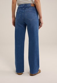 Person klädd i raka blåjeans och bruna sandaler, sedd bakifrån mot en plain ljus bakgrund.