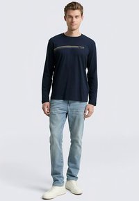 Navy long-sleeve t-shirt with yellow horizontal stitching, paired with light blue jeans and white sneakers. Smooth fabric, relaxed fit.