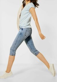 Donna che cammina, indossando una camicia a righe blu chiaro a maniche corte, jeans blu arrotolati, scarpe da ginnastica bianche, con una giacca leggera sulle spalle.