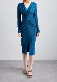 Teal glittrande klänning med långa ärmar, djup V-urringning och draperad front. Har en knä-lång slits och mjukt, texturerat tyg.