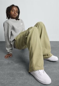 Barn med dreads iført en grå Nike-genser, vide beige bukser og hvite sko, sittende på et grått gulv mot en ensfarget vegg.