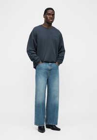 Mees, kellel on seljas tume, üle suuruse kampsun, laiad pleekinud sinised teksad ja mustad teravnurksed saapad, seisab käed taskutes.