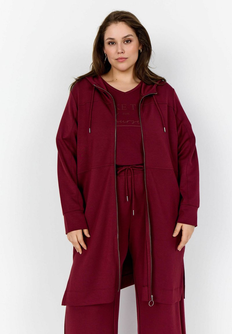 Burgunderfarbener Kapuzenpullover mit Reißverschluss, Kordelzug, langen Ärmeln und seitlichen Schlitzen, kombiniert mit einem passenden Oberteil. Glatte Textur und lockere Passform.