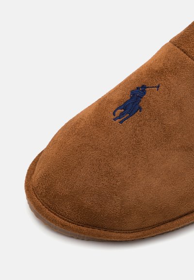 Bruine suède slof met een zachte textuur, voorzien van een blauwe geborduurde logo van een speler op een paard, gebogen vorm en gestikteDetails.