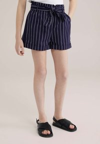 Jūras spēku svītrainas šortus ar sasietu jostu, kas aprīkoti ar elastīgu jostasvītru. Apvienoti ar melniem plakaniem sandaliem, kuru dizainā ir krusteniskas siksnas.