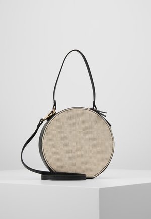 Sac à main rond en tissu beige avec garniture en cuir noir, poignée courte et bandoulière longue amovible, présenté sur un socle blanc.
