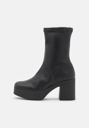 Bottines à talons noirs avec une texture lisse, bout rond et un talon bloc haut. Bord supérieur extensible pour un ajustement confortable.