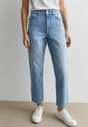 Person wearing high-waisted, light blue straight-leg jeans paired with white sneakers and a tucked-in white shirt, standing on beige floor.