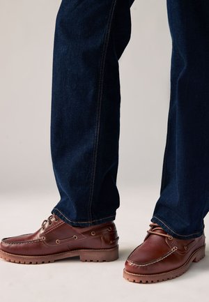 Jean bleu foncé porté avec des chaussures bateau en cuir marron dotées de semelles robustes et de détails à lacets.