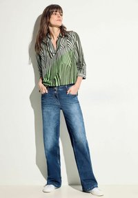 Blusa a righe verdi e nere con colletto, abbinata a jeans larghi blu scuro e sneakers bianche. Tessuto morbido con vestibilità rilassata.